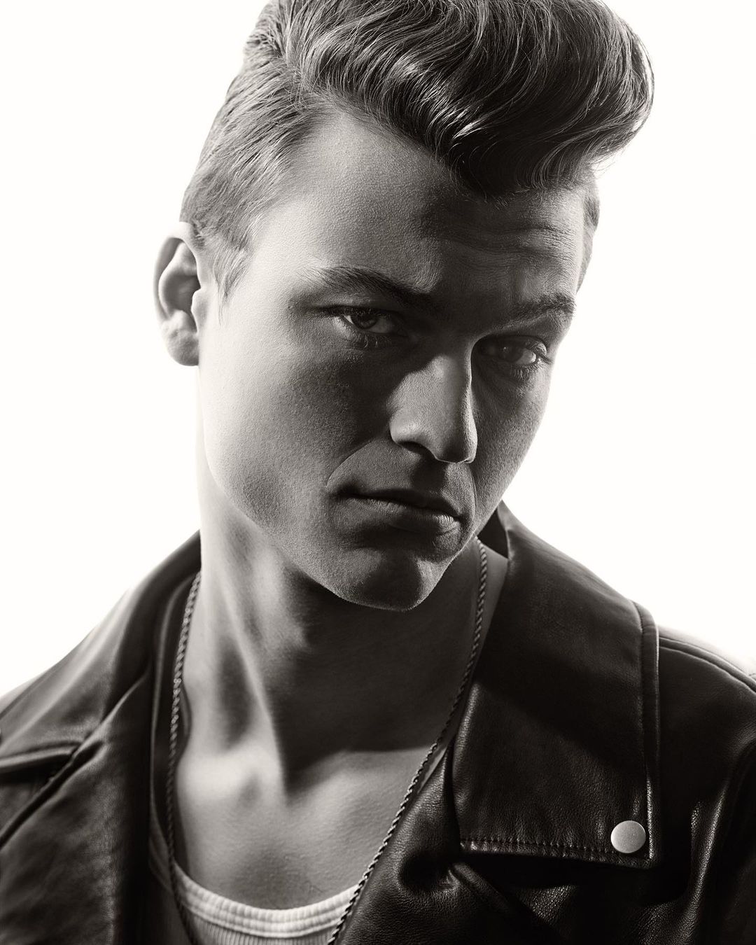 Monochrome portrait of a handsome adult male with pompadour hairstyle, wearing a leather jacket and chain necklace, against a white backdrop.